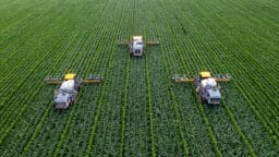 Three agricultural machines spray crops in parallel rows across a large green field, viewed from above.