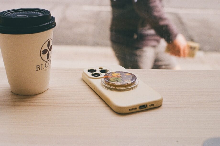 A coffee cup and a smartphone with a decorative pop socket sit on a light wooden table by a window, with a person walking outside.