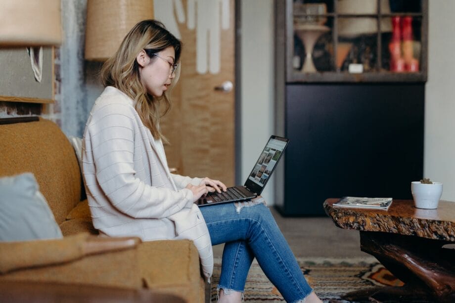 A woman sits on a sofa using a laptop, with a coffee table and a plant in a cozy indoor setting.