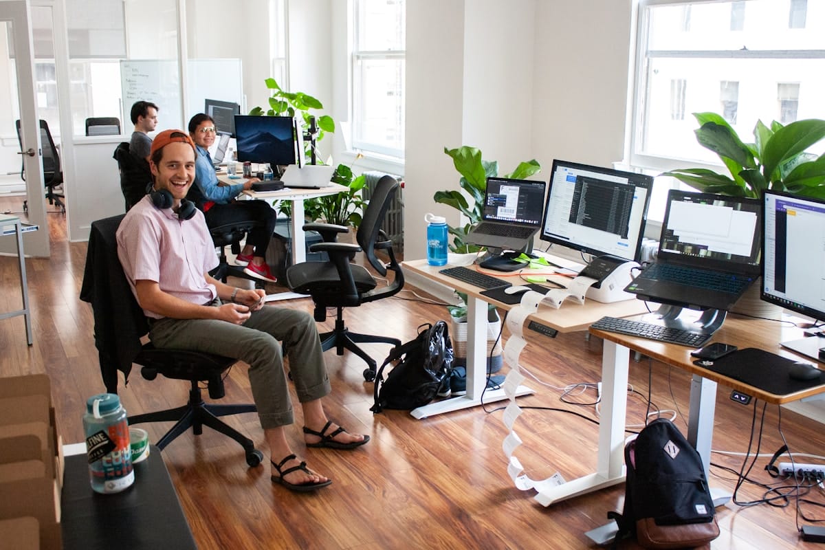 Four people work at desks with computers in a modern, bright office space with wooden floors and several green plants.