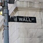 A Wall Street street sign attached to a metal pole in front of a stone building.