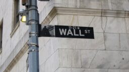 A Wall Street street sign attached to a metal pole in front of a stone building.