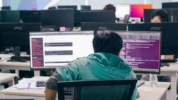 Person in a green jacket sits at a desk with two computer monitors, working in a modern, open-plan office with several other people.