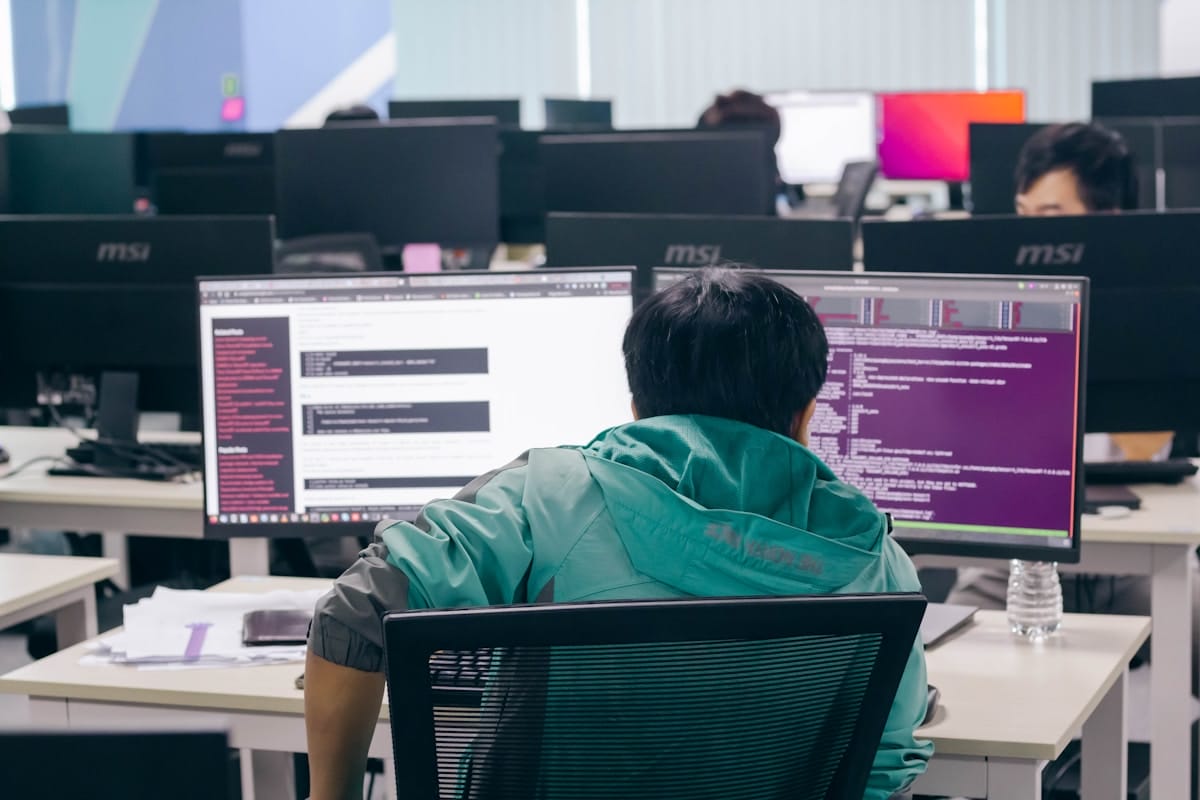 Person in a green jacket sits at a desk with two computer monitors, working in a modern, open-plan office with several other people.
