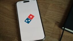 A smartphone displays the Dominos Pizza logo on its screen, resting on a wooden surface next to a black notebook.