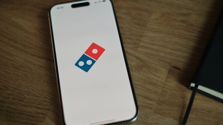 A smartphone displays the Dominos Pizza logo on its screen, resting on a wooden surface next to a black notebook.