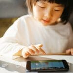 A young child in a white shirt plays a game on a smartphone at a table with a drink carton and small objects nearby.