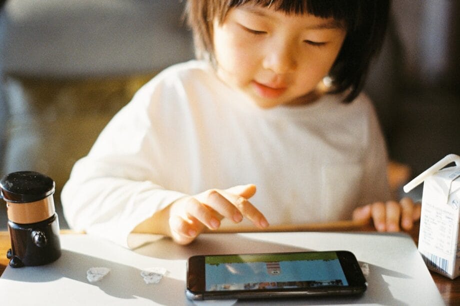 A young child in a white shirt plays a game on a smartphone at a table with a drink carton and small objects nearby.