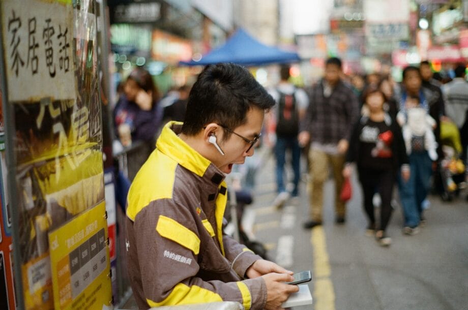 A man in a yellow jacket, wearing earphones, looks at his phone on a busy city street with blurred pedestrians in the background.