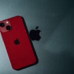 A red iPhone with dual cameras rests on a dark surface next to a black Apple logo.