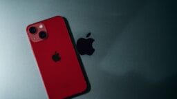A red iPhone with dual cameras rests on a dark surface next to a black Apple logo.