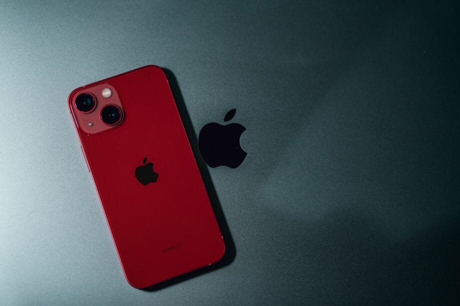 A red iPhone with dual cameras rests on a dark surface next to a black Apple logo.