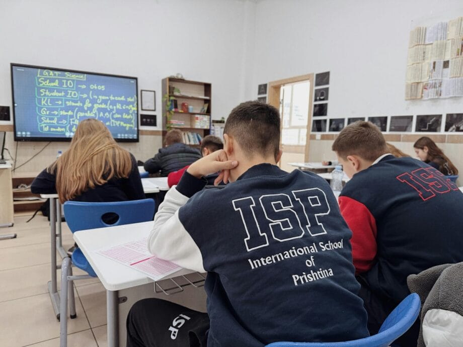 Students in ISP jackets sit at desks in a classroom, facing a digital whiteboard with notes displayed.