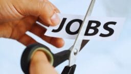 A hand uses scissors to cut a piece of paper with the word JOBS printed on it.