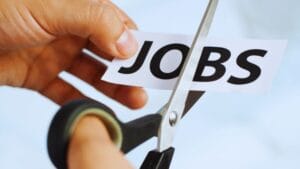 A hand uses scissors to cut a piece of paper labeled JOBS, symbolizing job cuts or layoffs.