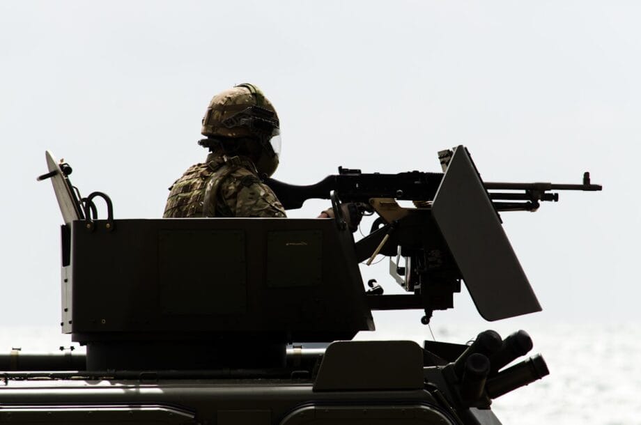 A soldier in tactical gear operates a mounted machine gun on a military vehicle against a bright, overexposed background.
