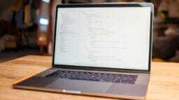 A laptop displaying code on its screen sits on a wooden table in a well-lit indoor room with blurred background.