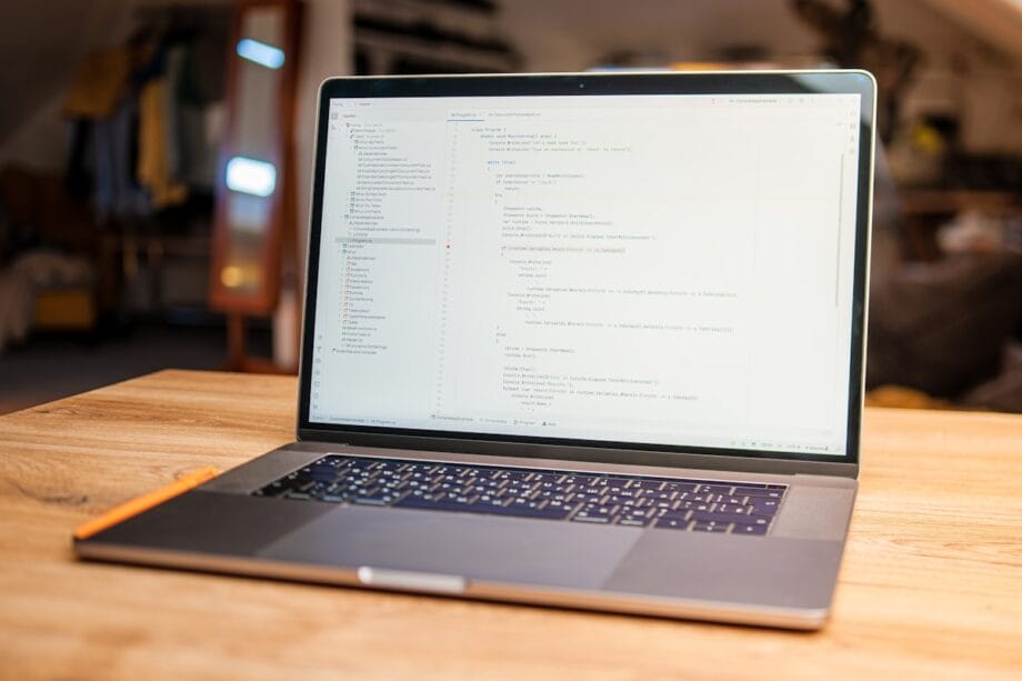 A laptop displaying code on its screen sits on a wooden table in a well-lit indoor room with blurred background.