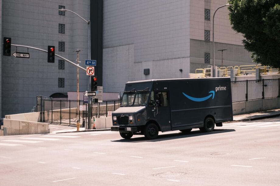 A dark blue Amazon Prime delivery truck drives through an empty city intersection on a sunny day.
