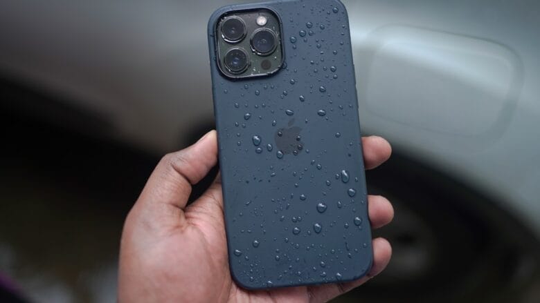 A hand holding a dark-colored iPhone with water droplets on its back, in front of a blurred car background.