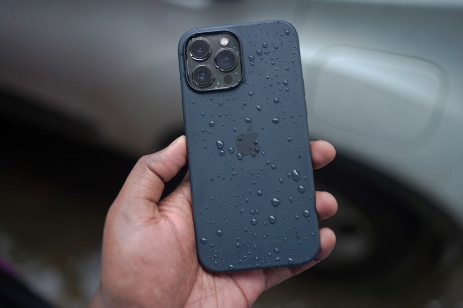 A hand holding a dark-colored iPhone with water droplets on its back, in front of a blurred car background.