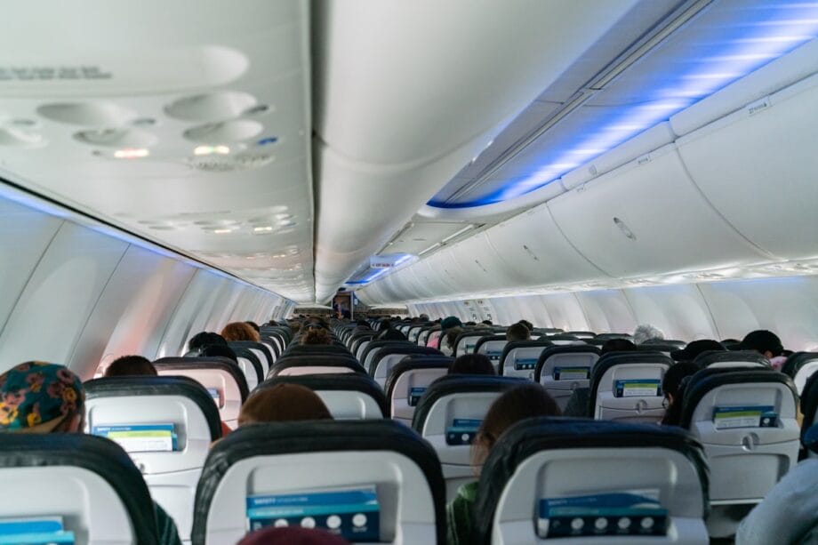 View from the back of an airplane cabin showing seated passengers and overhead bins with blue lighting.