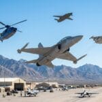 Several military aircraft, including jets and a helicopter, fly above an airbase with hangars and mountains in the background.