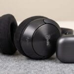 A pair of black over-ear headphones and a closed black earbuds case sit on a grey fabric surface.