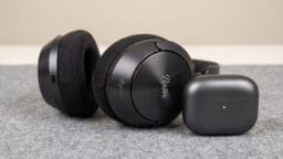 A pair of black over-ear headphones and a closed black earbuds case sit on a grey fabric surface.