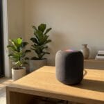 A smart speaker sits on a wooden table in a sunlit room with potted plants and stacked books in the background.