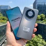 A person holds two Huawei Mate 70 Air smartphones, one showing the screen and the other showing the back, outdoors near glass buildings.