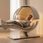 A man lies in a modern, pod-shaped relaxation chair in a bright, minimalist room with light wood flooring and neutral decor.