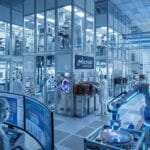 A high-tech semiconductor lab with engineers in cleanroom suits, robotic arms, computer monitors, and advanced manufacturing equipment.