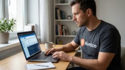 Man wearing a Kentico CMS shirt works on a laptop at a desk with a notepad, mug, and plant in a home office setting.