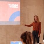 A woman presents a slideshow on digital evolution to a small group seated at a table with laptops in a modern meeting room.