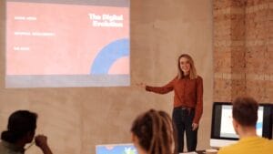 A woman presents a slideshow on digital evolution to a small group seated at a table with laptops in a modern meeting room.