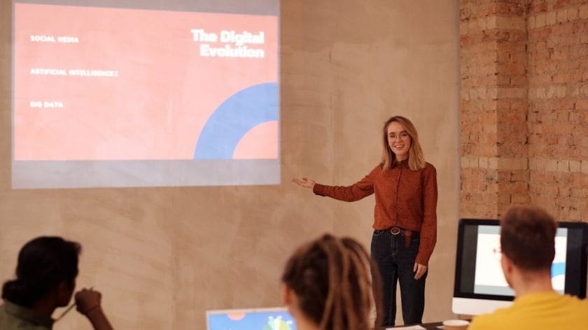A woman presents a slideshow on digital evolution to a small group seated at a table with laptops in a modern meeting room.