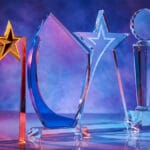 Four clear glass trophies with geometric shapes and star designs displayed against a blue and purple gradient background.