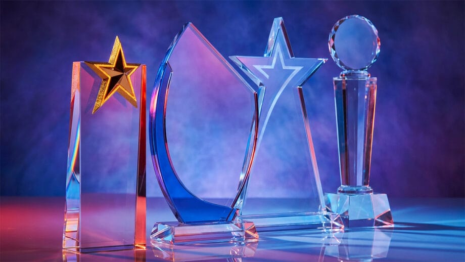 Four clear glass awards with geometric shapes and star designs displayed against a blue and purple gradient background.