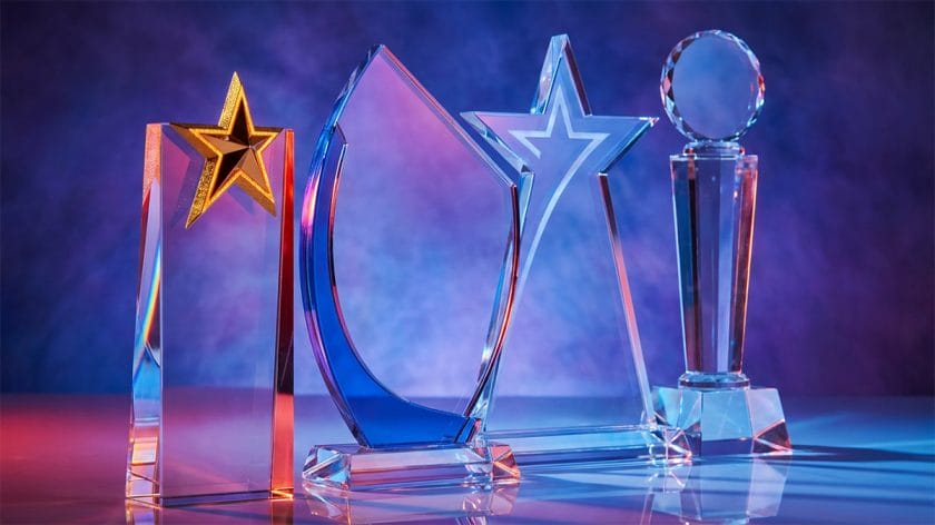 Four clear glass trophies with geometric shapes and star designs displayed against a blue and purple gradient background.