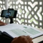 Person studying an open book while watching an online lecture on a smartphone, with a patterned screen in the background.