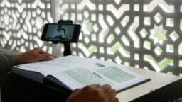 Person studying an open book while watching an online lecture on a smartphone, with a patterned screen in the background.