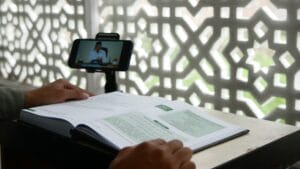 Person studying an open book while watching an online lecture on a smartphone, with a patterned screen in the background.