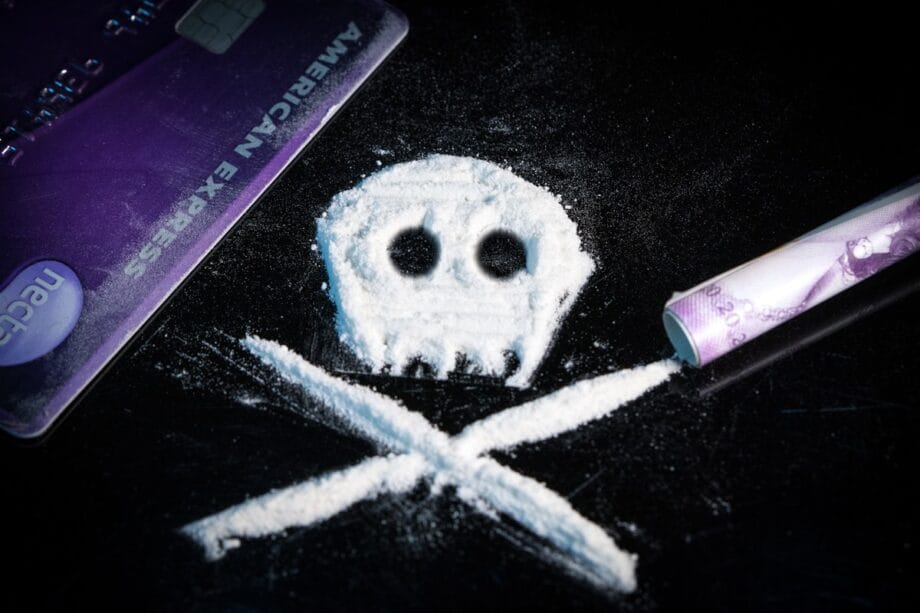 Lines of white powder arranged as a skull and crossbones, with a credit card and rolled banknote nearby on a dark surface.