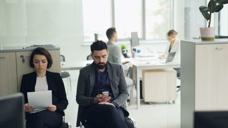 Two people sit in an office waiting area, one reading papers and the other using a phone, with coworkers visible in the background.