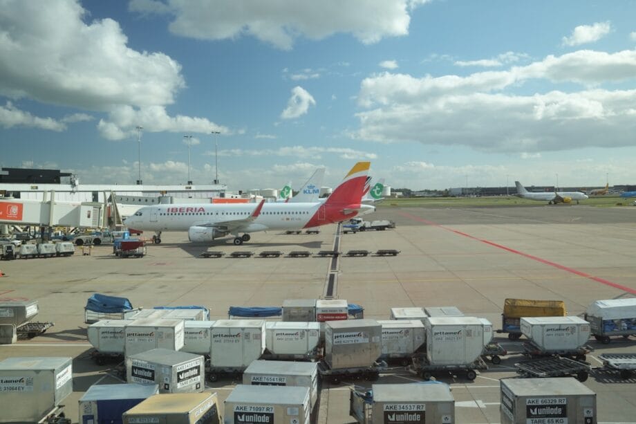 Passenger airplanes parked at airport gates with baggage carts and containers in the foreground under a partly cloudy sky.