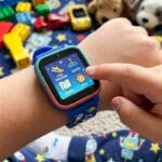A child’s hand uses a blue smartwatch displaying apps, with toys and building blocks scattered on a star-patterned surface in the background.