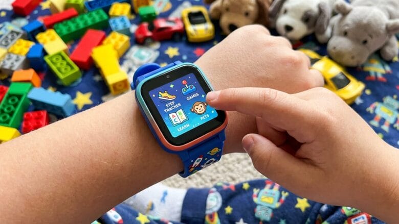 A child’s hand uses a blue smartwatch displaying apps, with toys and building blocks scattered on a star-patterned surface in the background.
