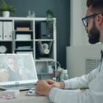 A doctor in a white coat sits at a desk, conducting a video call with a patient on a laptop in a modern office.