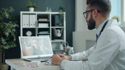 A doctor in a white coat sits at a desk, conducting a video call with a patient on a laptop in a modern office.
